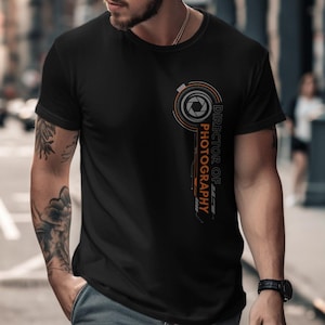 May include: Black crew neck t-shirt with a graphic design. The design features a camera lens with the words "Director of Photography" in orange and white. The shirt is made of a soft material.