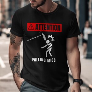 Può includere: Maglietta nera con un cartello di avvertimento rosso e bianco che dice "ATTENZIONE" e un omino bianco con un microfono che gli cade sulla testa. Il testo "FALLING MICS" è sotto l'omino.