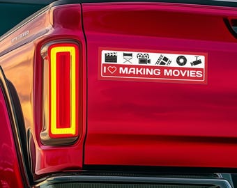 May include: A red truck with a white bumper sticker that says "I ♥ MAKING MOVIES" with icons of a clapperboard, director's chair, film reel, ladder, camera lens, and a microphone.