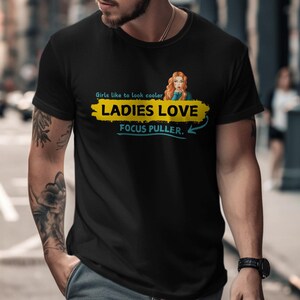 May include: Black t-shirt with a yellow and blue graphic that says "Girls like to look cooler. Ladies Love Focus Puller."