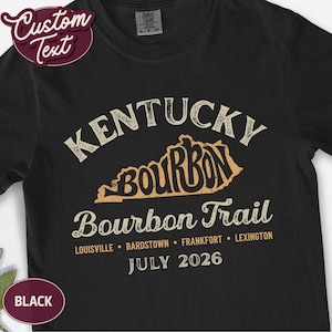 May include: A black short-sleeved t-shirt featuring a graphic with the text Kentucky Bourbon Trail, listing the cities Louisville, Bardstown, Frankfort, and Lexington, along with the date July 2026 and a map outline of the state of Kentucky.