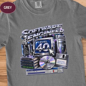May include: A grey t-shirt featuring a graphic print with the text "SOFTWARE ENGINEER" and "LEVEL 40". The design includes a computer monitor, a coffee mug labelled "CODE, COFFEE, COMMIT, REPEAT", a stack of books, a CD, a floppy disk, and a loading bar.