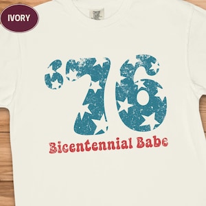 May include: Ivory-colored t-shirt with a distressed blue '76' graphic filled with white stars. The text 'Bicentennial Babe' is printed in red below the numbers. The shirt is made of a soft material.