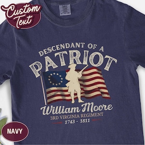 May include: A navy blue short-sleeve t-shirt featuring a graphic of a colonial soldier in front of an American flag. The text reads: DESCENDANT OF A PATRIOT, William Moore, 3RD VIRGINIA REGIMENT, 1743 - 1811. The design is printed in cream and red.