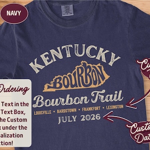 May include: A navy blue short-sleeve t-shirt featuring a graphic with the text Kentucky Bourbon Trail, a map outline, and city names including Louisville, Bardstown, Frankfort, and Lexington. The design includes the date July 2026 in white text.