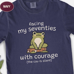 May include: Navy blue t-shirt featuring a cartoon frog sitting on a log, with the text "facing my seventies with courage (the cou is silent)". The shirt has a crew neck and short sleeves.