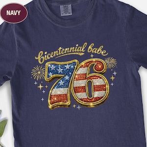 May include: Navy blue t-shirt with a gold glitter design that says "Bicentennial babe" above a sparkling "76" in red, white, and blue. Fireworks and stars complete the patriotic design.
