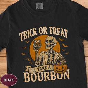 Può includere: Una t-shirt nera a maniche corte con la grafica di uno scheletro che tiene in mano un bicchiere di bourbon e un secchiello per caramelle a forma di zucca. Il testo recita TRICK OR TREAT e I'LL TAKE A BOURBON, su una luna arancione.