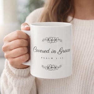 May include: White ceramic mug with the text "Covered in Grace" in a script font, with "Psalm 5:12" below. The mug features floral designs above and below the text. The mug is held by a person wearing a white sweater.