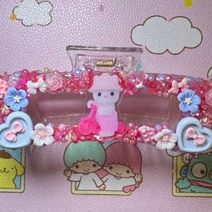 May include: A clear plastic hair clip decorated with pink and red beads, small bows, and flowers. The clip features a central figure of a pink bear with a pink accessory and heart-shaped accents. The background is a pink surface with cartoon characters.
