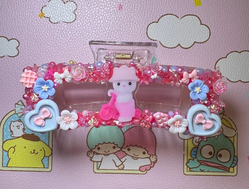 May include: A clear plastic hair clip decorated with pink and red beads, small bows, and flowers. The clip features a central figure of a pink bear with a pink accessory and heart-shaped accents. The background is a pink surface with cartoon characters.