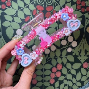 May include: A pink and clear hair claw clip decorated with pink beads, flowers, and a small pink figure. The clip features blue heart-shaped accents with bows and is held by a hand against a green floral background.