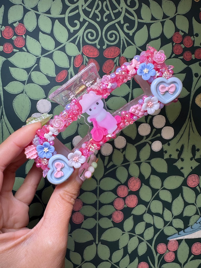 May include: A pink and clear hair claw clip decorated with pink beads, flowers, and a small pink figure. The clip features blue heart-shaped accents with bows and is held by a hand against a green floral background.