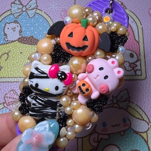 May include: A decorative Halloween-themed accessory featuring a pumpkin, a Hello Kitty figure wrapped in bandages, and a pink pig holding a pumpkin. The accessory is adorned with pearls and black accents, with a purple circular element at the top.