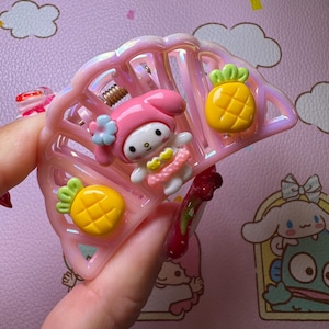 May include: Pink hair claw clip with a My Melody character and two yellow pineapple charms. The clip is shaped like a fan and has a spring mechanism. The background is a light pink color with cartoon characters.
