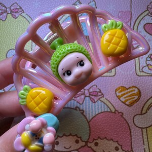 May include: A pink iridescent hair clip with a fan-like design. The clip is adorned with a smiling doll face wearing a green hat and two yellow pineapple charms. The background features a pink and white patterned design.