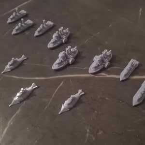 Puede incluir: Una colección de 12 barcos en miniatura de plástico gris, incluyendo submarinos y acorazados, dispuestos sobre una superficie gris.