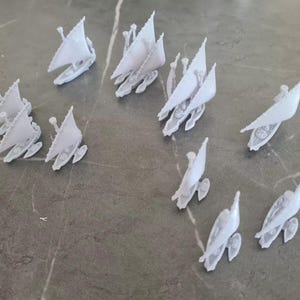 Puede incluir: Una colección de diez veleros en miniatura blancos, cada uno con una sola vela y un pequeño mástil. Los veleros están dispuestos de forma dispersa sobre una superficie gris claro.
