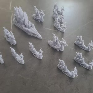Op de afbeelding: Een verzameling van 14 miniatuur drakenschipjes, geschilderd in een lichtgrijze kleur. De schepen zijn gemaakt van plastic en hebben ingewikkelde details, waaronder zeilen, masten en draken.