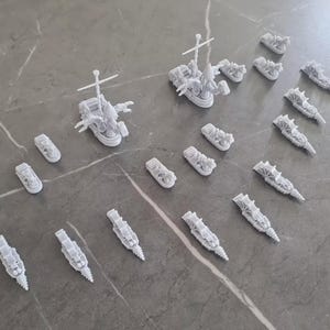 Puede incluir: Una colección de 20 barcos en miniatura blancos, incluyendo un barco más grande con un mástil elevado y una figura de proa. Los barcos están dispuestos sobre una superficie gris.