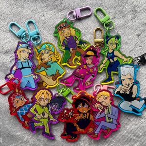 May include: A collection of ten acrylic keychains featuring various anime-style characters in colourful outfits, each attached to a plastic or metal lobster clasp. The characters display distinct hairstyles, clothing patterns, and poses, rendered in a vibrant, illustrated art style with clear outlines.