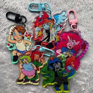 May include: Five acrylic keychains featuring colorful cartoon illustrations of various characters. The designs include a dizzy person, a winged figure with goggles, a pink-haired character with a blade, and two figures in dynamic poses with bright clothing.