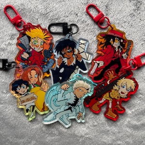 May include: A collection of five acrylic keychains featuring anime-style characters. The designs include figures in red, blue, and yellow outfits, some holding weapons or food items, attached to red or black plastic clasps for hanging.