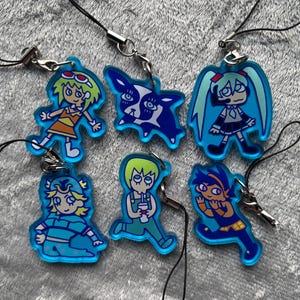May include: A collection of six translucent blue acrylic charms featuring various illustrated characters and a dog. Each charm is attached to a black cord with a silver-toned metal clasp, showcasing distinct graphic designs in vibrant colours.