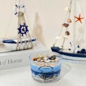 May include: A blue and white soy candle with seashells and a starfish inside. The candle is in a glass jar with a blue and white ocean-themed design. The text "Soy Candles" and "Farmina" is printed on the jar.