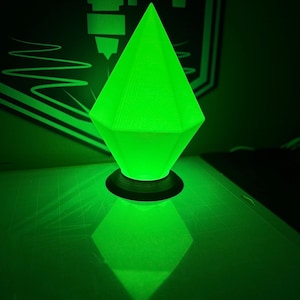Puede incluir: Una lámpara de forma de diamante, impresa en 3D, de color verde brillante con una base negra. La lámpara está encendida y proyecta un brillo verde.