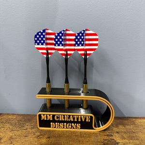 May include: A black dart holder with a gold trim and the text "MM CREATIVE DESIGNS". Three darts with American flag designs are in the holder.
