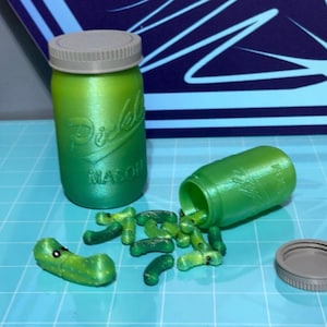 Op de afbeelding: Een groene glazen pot met een grijs deksel, met het label "Pickle Mason", laat groene, komkommerachtige voorwerpen eruit vallen.