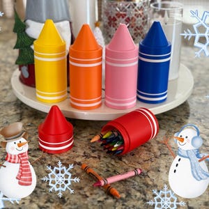 Puede incluir: Un conjunto de seis recipientes de crayones de colores, cada uno con forma de crayón. Los recipientes están dispuestos en una fila sobre una superficie blanca. Los recipientes son rojo, naranja, amarillo, verde, azul y morado. También hay dos figuras de muñeco de nieve en la superficie.