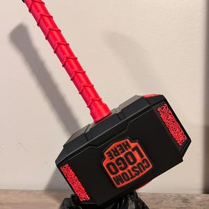 May include: A black and red prop hammer with the text "CUSTOM LOGO HERE". The hammer has a red handle and decorative red accents. It rests on a black, textured base.