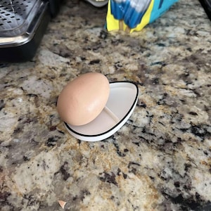 May include: A white egg holder with a black rim, shaped like a teardrop, with a single brown egg inside.