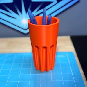May include: An orange 3D-printed pen holder with vertical grooves and blue pens inside. The pen holder is cylindrical and sits on a blue grid-patterned surface. The background features a blue geometric design.