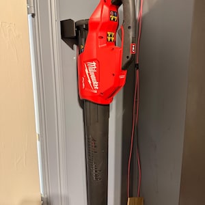 May include: A red and black Milwaukee FUEL cordless leaf blower hanging on a gray wall. The blower has a black nozzle and handle, with the Milwaukee logo visible. The handle has several buttons and indicator lights.