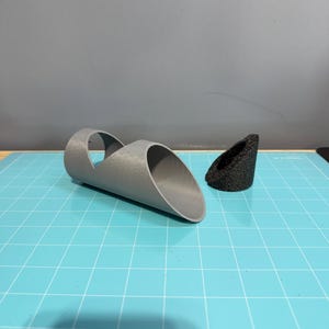 Può includere: Un oggetto grigio stampato in 3D con due fori, e un oggetto più piccolo di colore nero stampato in 3D con un foro. Entrambi gli oggetti sono su una superficie a griglia blu.