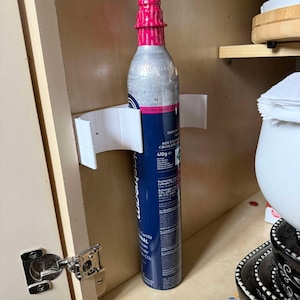 May include: A white plastic holder for a silver and blue carbon dioxide canister. The canister is labeled "SodaStream" and has a pink cap. The holder is mounted on a wooden cabinet door.