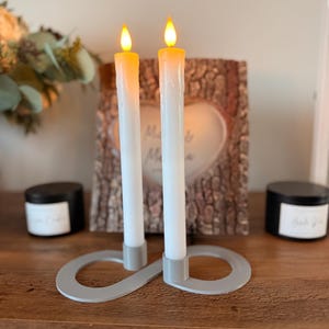 May include: Two white pillar candles with a warm yellow flame are displayed on a gray infinity-shaped candle holder. The candles are on a wooden surface.