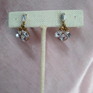 May include: A pair of gold earrings with clear crystal stones. The earrings are on a white display stand.
