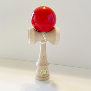 May include: A red wooden kendama with a natural wood base and a string connecting the ball to the base. The base has a small gold sticker with the text "KENDAMA" on it.
