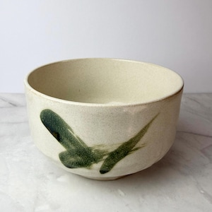 May include: A cream-colored ceramic bowl with a speckled texture. The bowl features a dark green brushstroke design on the exterior. The bowl has a wide, open shape, suitable for serving or display.