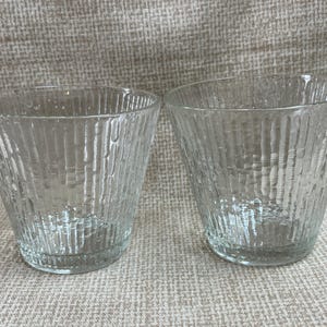 May include: Two clear glass tumblers with a textured, ribbed design. The glasses are empty and sitting on a textured surface.