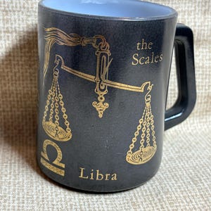 May include: Black ceramic mug featuring a gold Libra zodiac design. The mug has a black handle and a light blue interior. The design includes the Libra symbol, scales, and the words "the Scales" and "Libra".