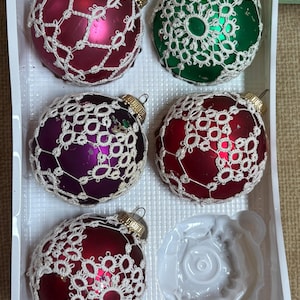 May include: A set of five Christmas ornaments in a white plastic tray. The ornaments are glass spheres in shades of red, green, and purple, each covered with a white lace overlay. Gold-colored metal caps are at the top of each ornament.