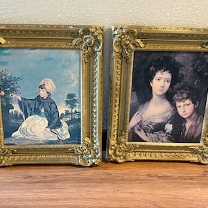 May include: Two rectangular wall art pieces in ornate gold frames. The left print depicts a child in a white dress and dark cape near a bush. The right print shows a portrait of an adult and a child with dark hair, set against a dark background.