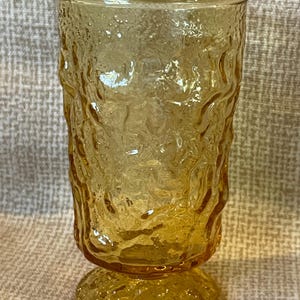 May include: A textured, amber-colored glass with a pedestal base. The glass has a rough, organic pattern on its surface, giving it a unique, handcrafted appearance. The stemware is suitable for serving beverages.