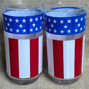 May include: Two clear drinking glasses with a patriotic design. The top section features a blue band with white stars, while the lower section has red and white stripes. These glasses are perfect for celebrating Independence Day.