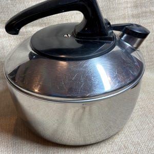 May include: A stainless steel tea kettle with a black handle and lid. The kettle has a rounded body and a spout with a black cap. A white button is on the handle. The kettle is designed for stovetop use.